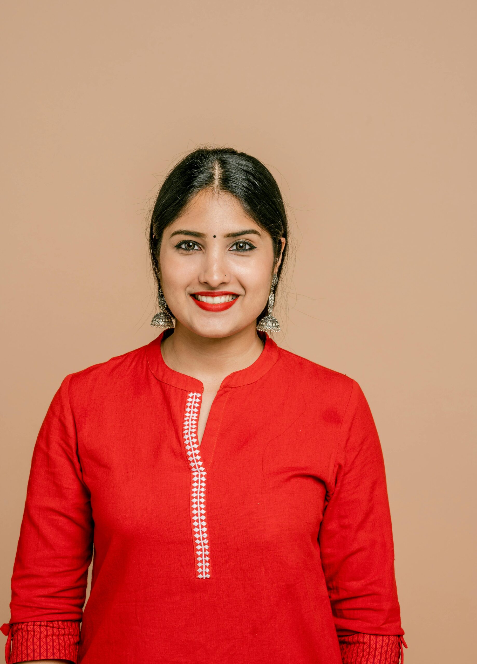 Portrait of a smiling woman wearing a red dress, earrings, and lipstick against a beige background.